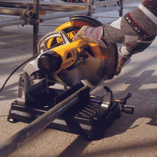 A person wearing gloves is operating a DEWALT power saw, cutting a metal pipe. The saw is positioned on a flat surface outdoors, with construction scaffolding visible in the background.