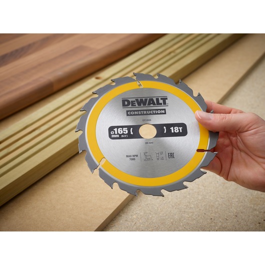 Hand holding a DEWALT construction circular saw blade, model DT1933, with 165 mm diameter and 18 teeth, in front of wooden planks and boards.