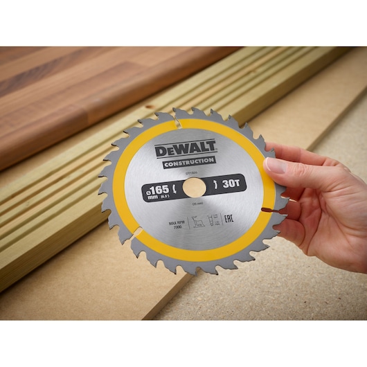 A hand holding a DEWALT Construction 165 mm 30-tooth circular saw blade, model DT1935, with wood planks in the background.