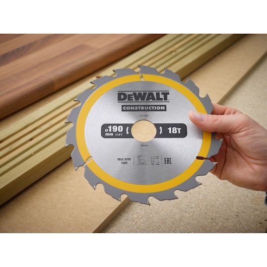 A hand holding a DEWALT Construction circular saw blade, model DT1943, with a diameter of 190 mm (7.5 inches) and 18 teeth. The blade is shown in front of wooden boards on a workbench.
