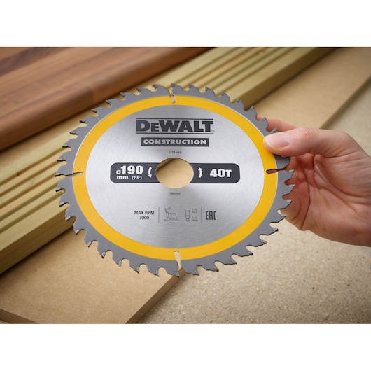 A hand holding a circular saw blade with a yellow ring, branded DEWALT Construction, model DT1945. The blade is marked 190 mm (7.5 inches) and 40 teeth, designed for cutting wood. Wooden boards are visible in the background.
