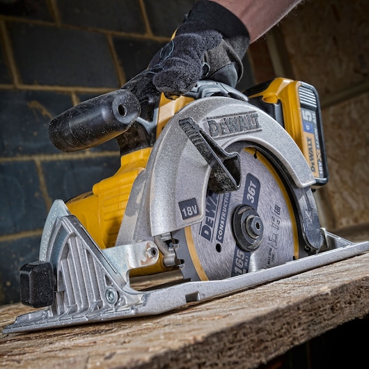 A close-up view of a DEWALT DT1950 cordless circular saw being used to cut a piece of wood. The saw is held by a person wearing a black glove, and the tool displays its brand and technical details, such as 18V power and 165mm blade size.