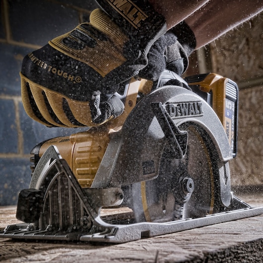 Close-up of a gloved hand operating a DEWALT cordless circular saw cutting through wood, with sawdust particles visible around the tool.