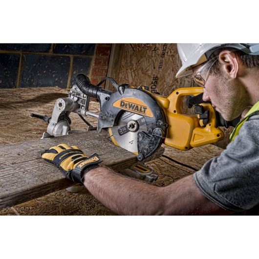 A person wearing safety gloves and a helmet is using a DEWALT circular saw to cut a wooden plank in a workshop or construction setting.