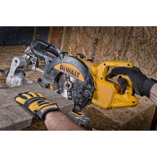 A person wearing protective gloves is using a DEWALT circular saw to cut a piece of wood on a construction site. The saw is held securely and dust extraction equipment is attached.