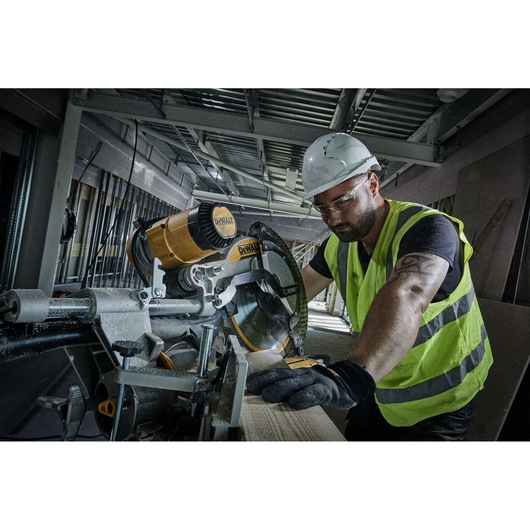 A person wearing a safety helmet, gloves, and a high-visibility vest is cutting wood with a DEWALT miter saw in an industrial construction setting.