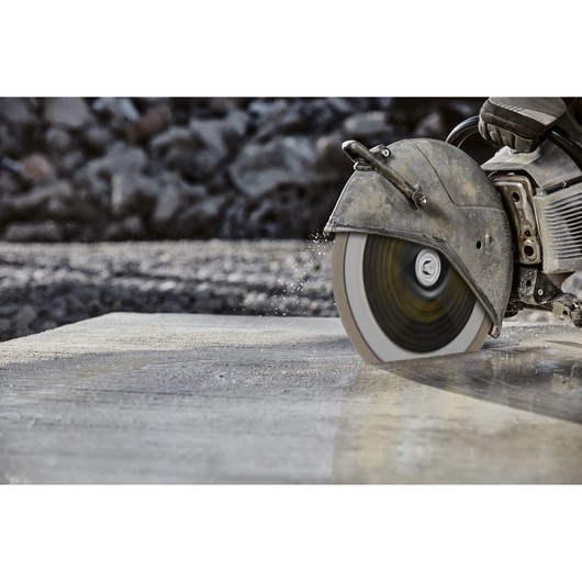 A close-up of a power saw cutting through wet concrete outdoors, water splashing from the blade.