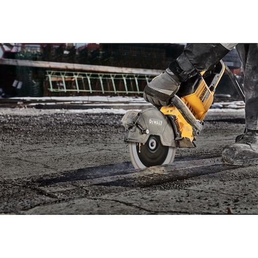 A DEWALT power saw being used to cut through pavement at a construction site. A person is operating the saw, wearing protective gloves and boots.