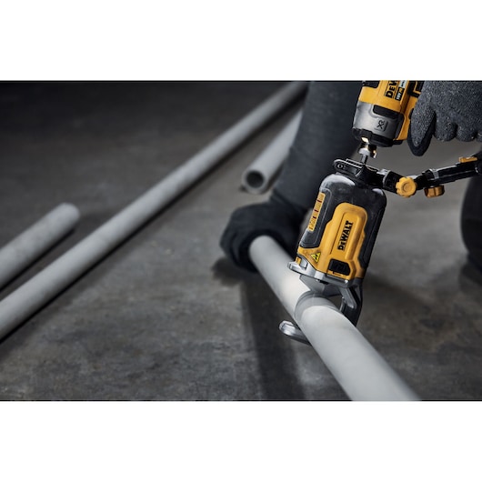 Close-up of a DEWALT power tool being used by a person wearing gloves to cut a grey metal pipe on a workshop floor.