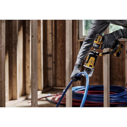 A person wearing gloves uses a DEWALT DT20560 power tool to cut a blue plastic pipe in a wooden-framed construction interior.