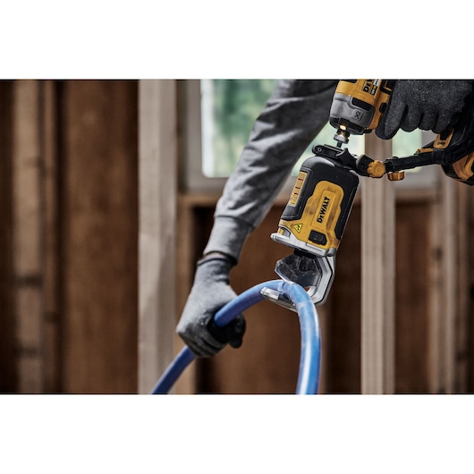 A person wearing gloves uses a DEWALT DT20560 power tool to cut a blue plastic pipe in a construction setting.