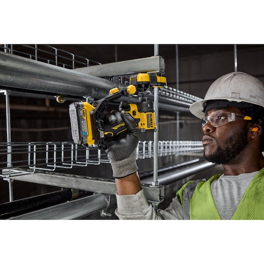 Worker uses DEWALT cordless drill to install metal conduit, wearing gloves, safety vest, and helmet.