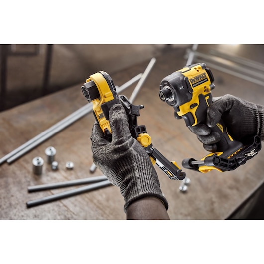 Gloved hands hold a DEWALT impact driver and accessory in a workshop, with metal rods and parts in the background.