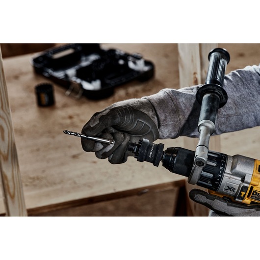 A close-up of a person wearing work gloves inserting a drill bit into a DEWALT power drill, with a blurred background of tools and wood.
