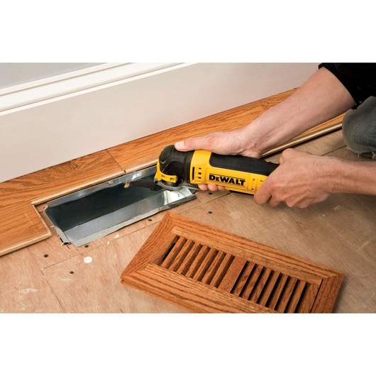A person uses a yellow and black DEWALT power tool to cut wood flooring near a metal vent opening. A wooden vent cover is placed nearby on the floor.