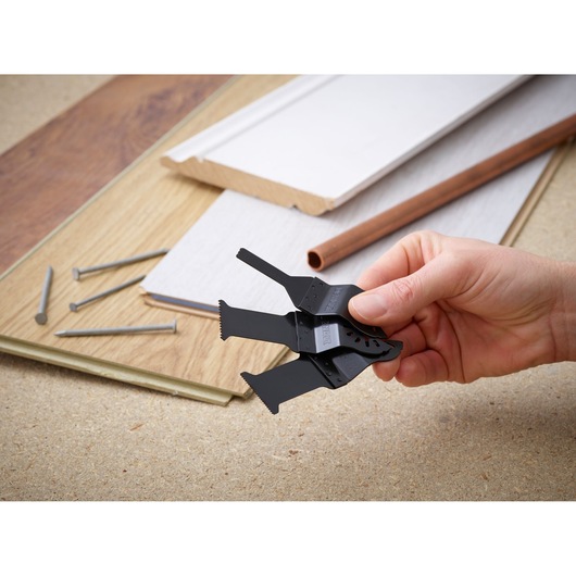 A hand holds several DEWALT DT20713 multi-tool blades, with woodworking materials, nails, and a pipe in the background.