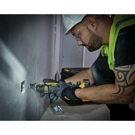A person wearing safety gear uses a DEWALT oscillating multi-tool to cut into a wall, with screws and a metal outlet box on the floor nearby.