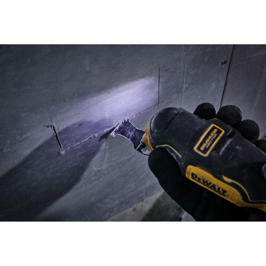 A DEWALT multi-tool cutting a rectangular hole in a drywall panel, held by a person wearing black gloves. The tool is illuminated by a built-in light.