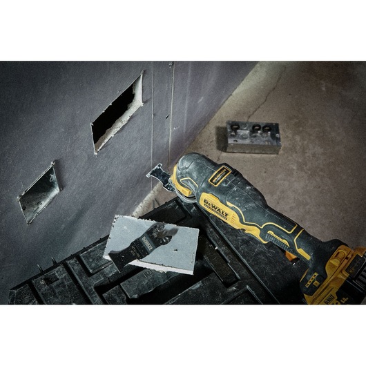 A DEWALT oscillating multi-tool being used to cut a rectangular hole in drywall, with the cutout piece and the tool placed on a black toolbox. There are visible cut sections on the wall and construction materials in the background.