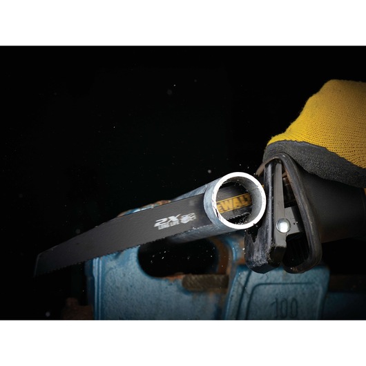 A close-up view of a DEWALT reciprocating saw cutting through a metal pipe, held by a gloved hand. The saw blade is actively slicing the pipe, with metal shavings visible.