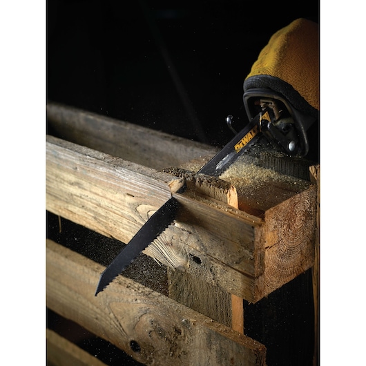 A DEWALT reciprocating saw blade attached to a power tool is cutting through a wooden pallet, with wood dust visible around the cut.