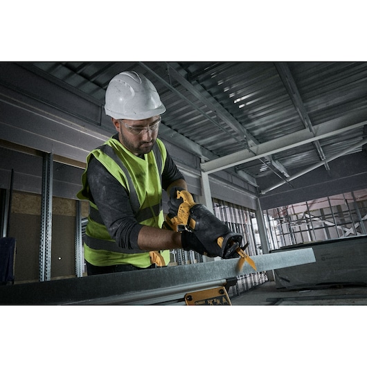 A worker wearing a hard hat, safety vest, and gloves uses a yellow DEWALT reciprocating saw to cut through a metal beam inside a construction site.