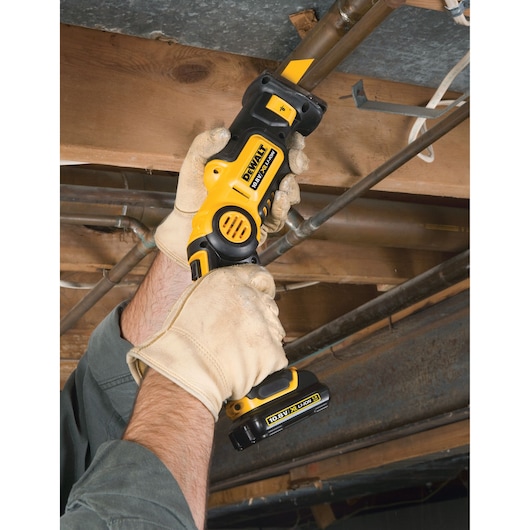 Gloved hands using a DEWALT cordless reciprocating saw to cut a pipe in an unfinished ceiling area.