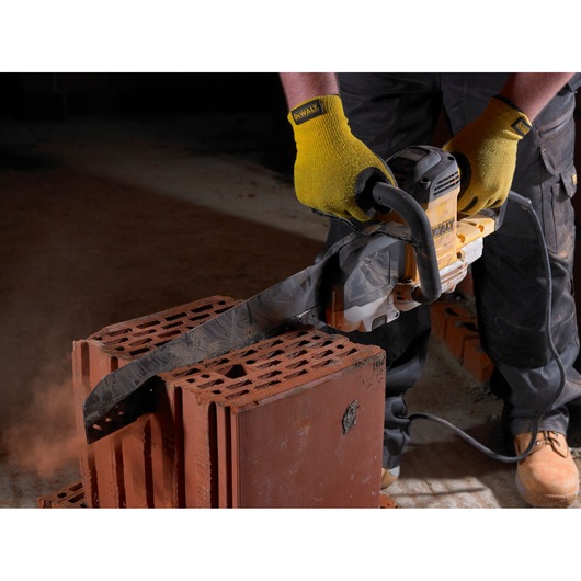 A person wearing yellow gloves uses a DEWALT power saw to cut through red construction bricks. The saw creates dust as it slices through the bricks, which are stacked on the ground.