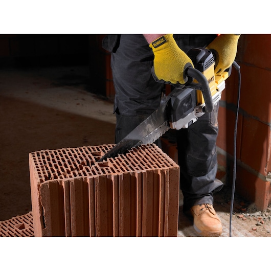 Person uses a DEWALT power saw to cut a hollow brick block indoors. Hands wear yellow gloves and boots are visible.