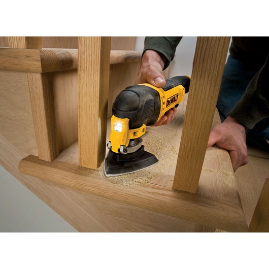 A person uses a DEWALT detail sander to sand wooden stairs, focusing on tight corners and edges, with wood dust visible around the tool.