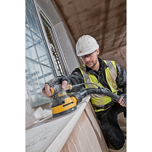 Person using a DEWALT sander on a windowsill, wearing safety gear and high-visibility vest at a construction site.