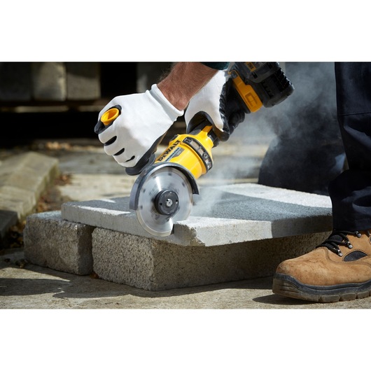 A person wearing work gloves uses a DEWALT angle grinder to cut a concrete block, creating dust. The scene shows protective footwear and the tool in action.