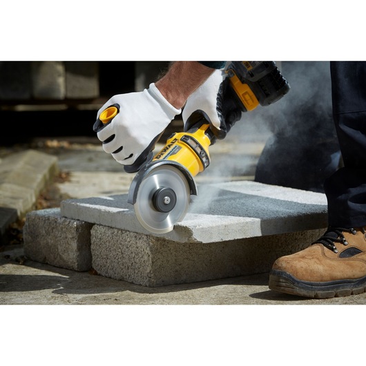 A person wearing white gloves uses a yellow DEWALT power tool to cut through a concrete slab, with dust rising from the cutting area. The scene is outdoors, and one person is wearing brown work boots.