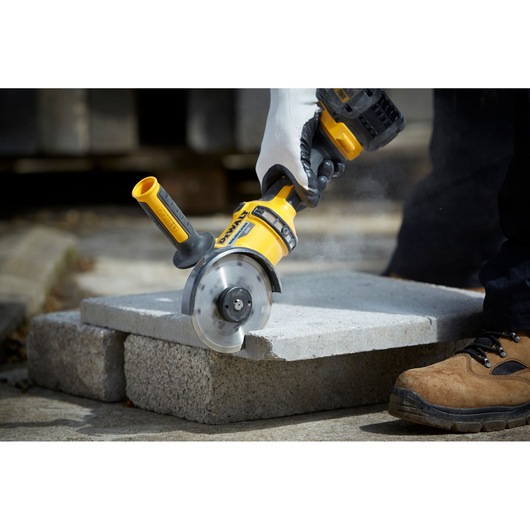 A DEWALT hand-held power tool cutting through a concrete slab, operated by a person wearing gloves and work boots.