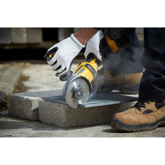 A person wearing work gloves uses a yellow DEWALT power tool to cut a slab, producing dust. The person is also wearing sturdy work boots and working outdoors.