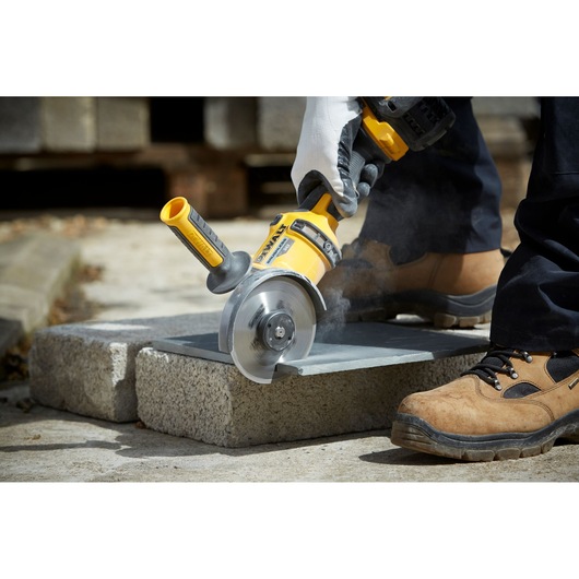 A person wearing work gloves and safety boots is using a yellow DEWALT power tool with a circular blade to cut a stone slab outdoors. Dust is visible as the tool operates.