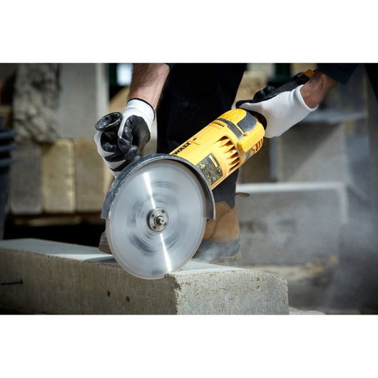 A person wearing gloves is using a yellow DEWALT power tool with a circular blade to cut through a concrete block outdoors.