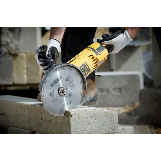 A close-up view of a person using a yellow DEWALT power tool with a circular blade to cut through a concrete block. The individual is wearing protective gloves.