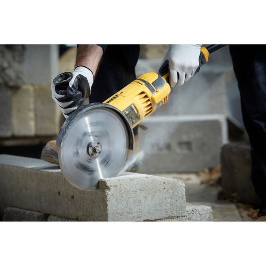 A person wearing gloves is using a yellow DEWALT angle grinder to cut a concrete block. The tool is producing dust as it operates.