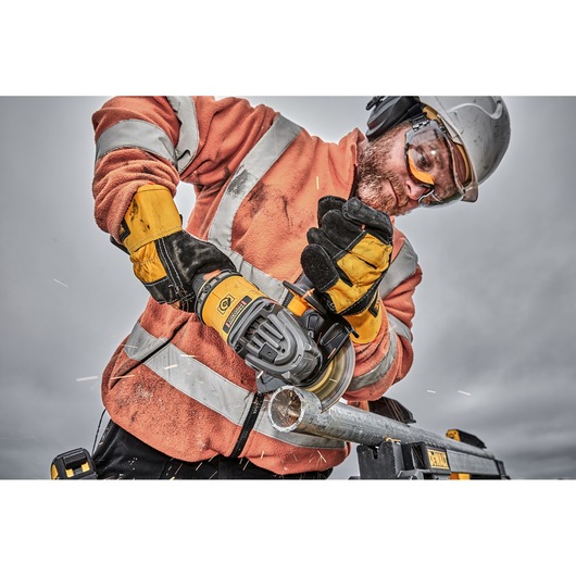 A person wearing an orange high-visibility jacket, yellow gloves, and a safety helmet is using a DEWALT angle grinder to cut a metal pipe. Sparks are visible around the cutting area. The person's face is obscured for privacy.