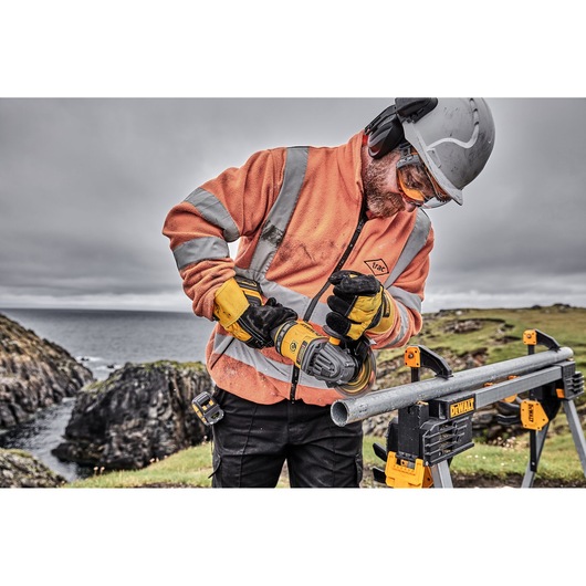 A person wearing safety gear, including a helmet, gloves, and an orange high-visibility jacket, is using a DEWALT power tool to grind or cut a metal pipe outdoors. The pipe is secured on a DEWALT stand, with rocky terrain and water visible in the background.