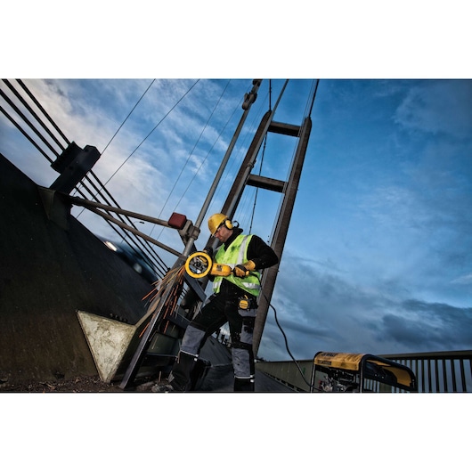 Worker uses a DEWALT grinder on metal near a bridge, wearing safety gear and high-visibility vest.