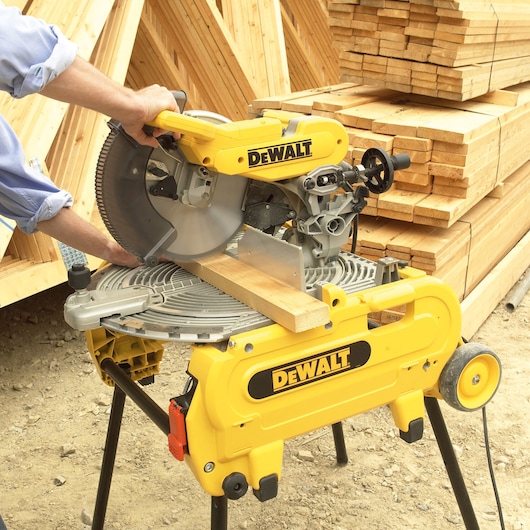 A person is using a yellow DEWALT miter saw to cut a wooden plank at a construction site, with stacks of lumber in the background.