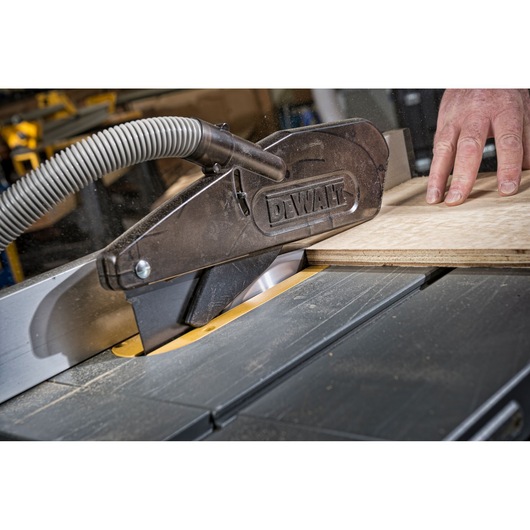 Close-up of a DEWALT table saw in use, with a dust extraction hose attached and a hand guiding a piece of plywood through the saw.