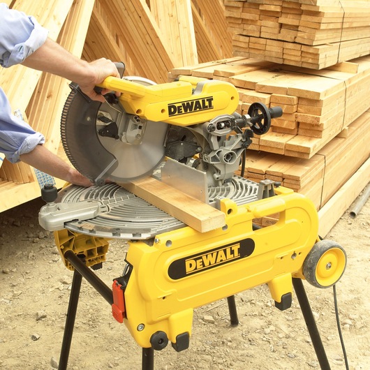 A DEWALT miter saw is being used to cut a piece of wood on a construction site, with stacks of lumber in the background.