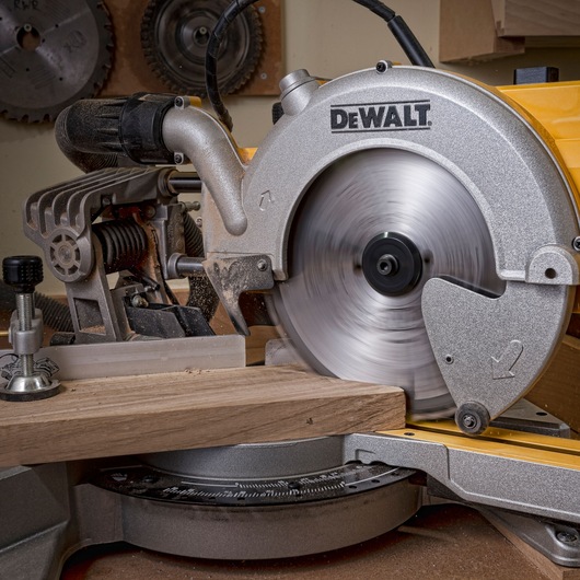 A close-up view of a DEWALT branded miter saw cutting through a piece of wood, with the spinning blade in motion, in a workshop setting.