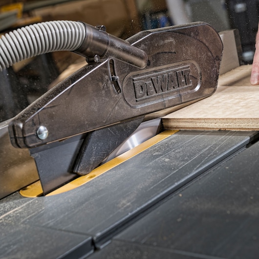 Close-up view of a DEWALT table saw in use, cutting a sheet of wood. The saw blade is partially covered by a safety guard with a dust extraction hose attached.