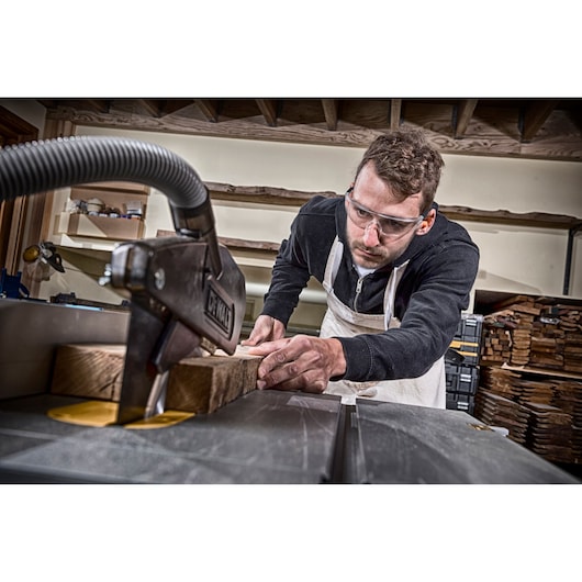 A person wearing a black sweatshirt and white apron operates a DEWALT table saw to cut a piece of wood in a workshop environment. The focus is on the woodworking process, with stacks of wood and tools visible in the background.
