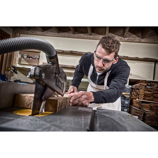 A person wearing a black sweatshirt and apron is operating a DEWALT table saw to cut a piece of wood in a woodworking workshop. The background shows shelves with tools and stacked wood.
