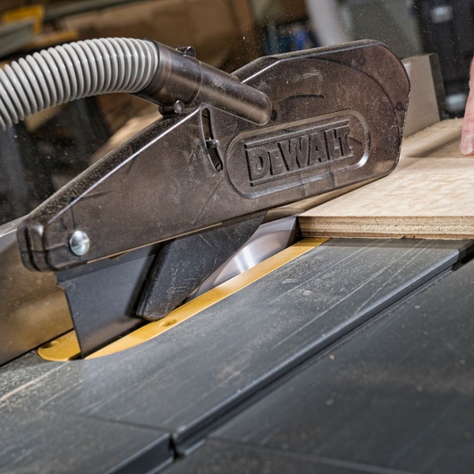 Close-up of a DEWALT table saw with a dust extraction attachment, cutting a piece of plywood.